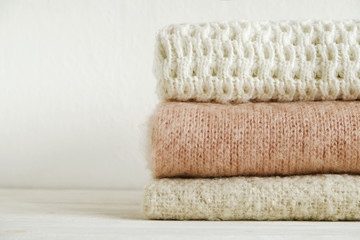 Fototapeta na wymiar Bunch of knitted warm pastel color sweaters with different knitting patterns folded in stack on white wooden table, textured wall background. Fall winter season knitwear. Close up, copy space for text