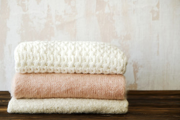 Fototapeta na wymiar Bunch of knitted warm pastel color sweaters with different knitting patterns folded in stack on brown wooden table, white textured wall background. Fall winter knitwear. Close up, copy space for text.