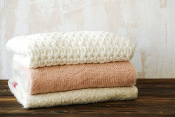 Obraz na płótnie Canvas Bunch of knitted warm pastel color sweaters with different knitting patterns folded in stack on brown wooden table, white textured wall background. Fall winter knitwear. Close up, copy space for text.