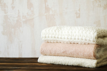 Fototapeta na wymiar Bunch of knitted warm pastel color sweaters with different knitting patterns folded in stack on brown wooden table, white textured wall background. Fall winter knitwear. Close up, copy space for text.