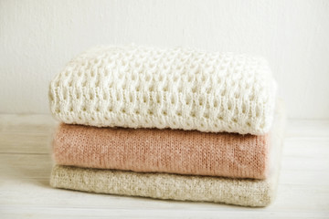 Obraz na płótnie Canvas Bunch of knitted warm pastel color sweaters with different knitting patterns folded in stack on white wooden table, textured wall background. Fall winter season knitwear. Close up, copy space for text
