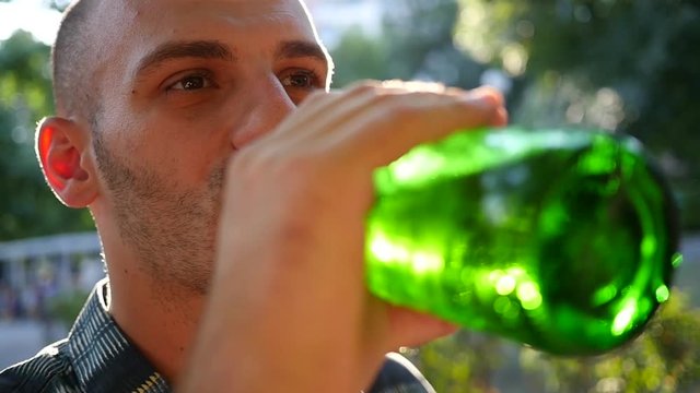Man drinking alcohol beer from bottle outdoors in park in sun back light haze slow motion