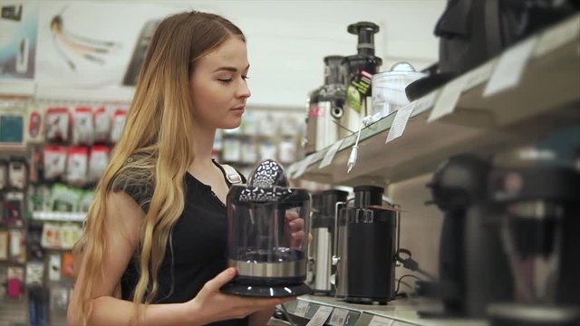 Pretty girl is standing in a big store alone and holding small electrical coffee-machine in hands. She is want to buy and looking on other models on a shelf