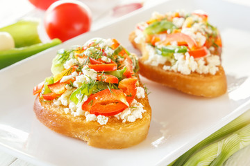 Two Bruschetta with red sweet pepper and goat cheese in a plate next to fresh vegetables on white boards. Close-up