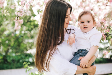 A happy mother with a baby in her arms in white clothes looks into his eyes against background of cherry blossom