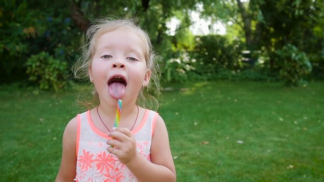 Little child girl eat lick tongue a sweet colored caramel candy stick in green garden
