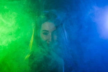 Portrait of young woman in neon blue and green smoke with vape or e-cigarettes