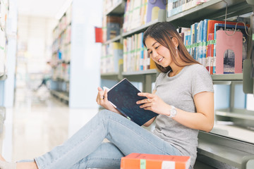 Asian Woman reading and sitting near a bookshelf in the library..
