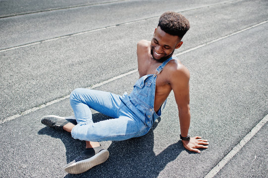 Handsome sexy african american bare torso man at jeans overalls sitting at stadium racecourse. Fashionable black man portrait.