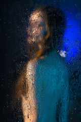 Blurred silhouette of a beautiful slender naked girl standing behind glass covered with drops of...