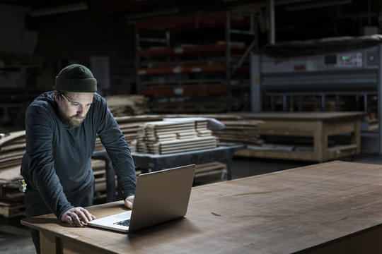 A Caucasian carpenter working on his lap top after hours in a large woodworking factory.