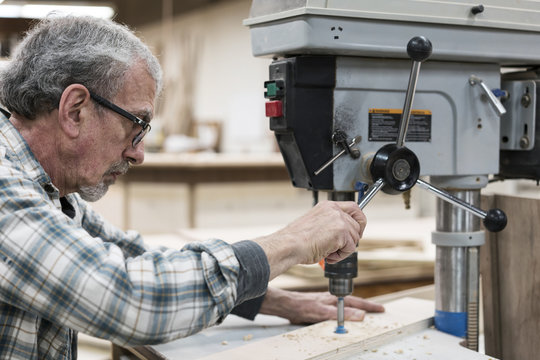 A senior man with glasses and beard in a woodworkers shop, using a machine. 