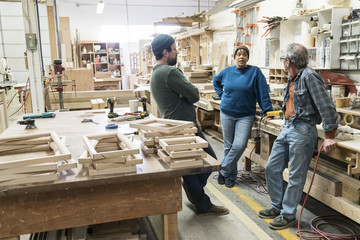 A group of mixed race carpenters discussing a project at a work station in a large woodworking shop.