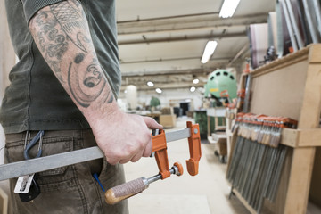 Closeup of an arm with tatoos and a hand holding a bar clamp in a large woodworking factory.