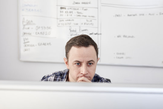 Man looking at a computer screen seated at his desk, with a project chart on the whiteboard behind.