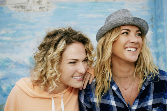 Portrait of two smiling women with blond hair wearing pink hoodie, blue checkered shirt and grey Trilby hat.