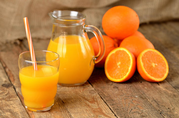 Plakat Glass with orange juice and straw, jug with fresh juice and pile of oranges in the background on wooden table