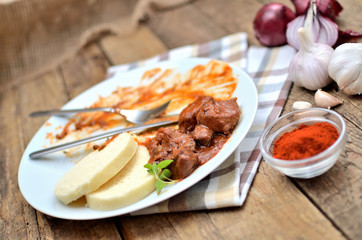 Eating pork goulash meat with dumplings on white plate, cutlery, garlic, onion, pepper, tablecloth in the background - typical Czech food