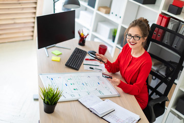 A young girl is sitting at the desk in the office, holding a bank card and phone in her hand.