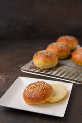 Homemade Hamburger Buns Topped with Sesame Seeds on a Rustic Black Background; One Cut Open on White Square Paper Plate
