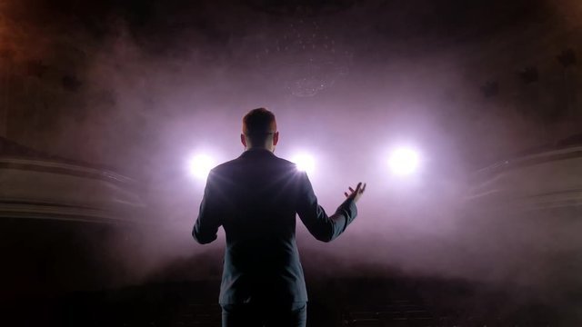 Showman. Young male entertainer, presenter or actor on stage. Back, arms to sides, smoke on background of spotlight. Rear view of a male public speaker speaking at the microphone, pointing, in the