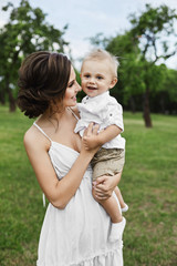 Fototapeta premium Beautiful and sensual brunette model girl in short white dress, with cute little stylish baby boy on her hands posing at green park