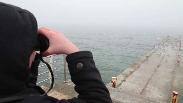 Closeup portrait of young kid looking through black vintage binoculars at something far away in distance at cloudy sea.