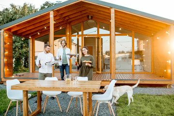 Happy friends carrying food preparing for the dinner on the backyard of the modern wooden house during the sunset