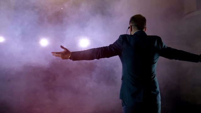 Showman. Young male entertainer, presenter or actor on stage. Back, arms to sides, smoke on background of spotlight. Rear view of a male public speaker speaking at the microphone, pointing, in the