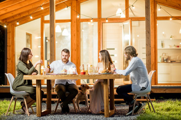 Friends having a dinner sitting at the table on the backyard of the modern house decorated with lights during the evening time outdoors