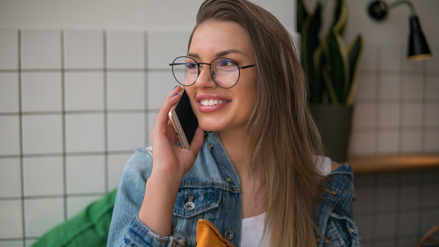 Beautiful young woman in glasses, speaks on the phone, has pleasant smile, dressed in jeans shirt, indoors cafe