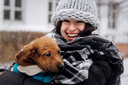 stylish hipster woman hugging and smiling cute puppy in snowy cold winter park. moments of true happiness. adoption concept. save animals
