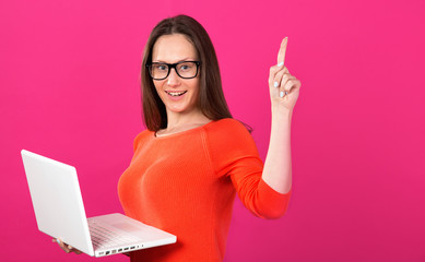 Beautiful brunette woman with glasses. Young woman 25 years old with a laptop in her hands showing a finger up standing on a pink background. Online shopping.