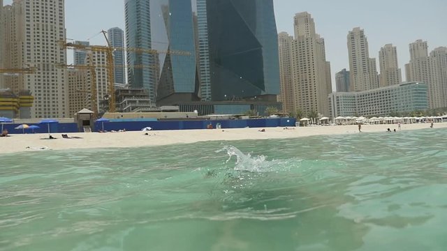An optimistic view of a cheerful small boy jumping and diving feet first in the light green waves on a sandy beach in Dubai in summer in slo-mo