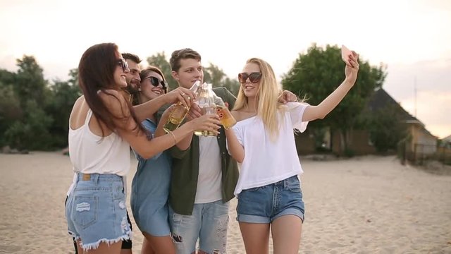 Group of friends having fun enjoying a beverage and relaxing on the beach at sunset in slow motion. Young men and women drink beer standing on a sand in the warm summer evening.
