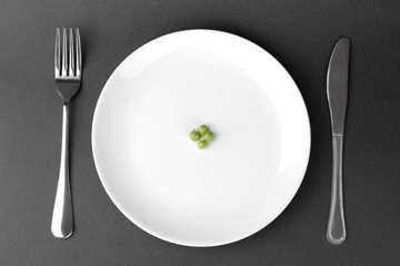 a few green fresh peas on a white plate next to a fork and knife on a black background. The concept of diet.