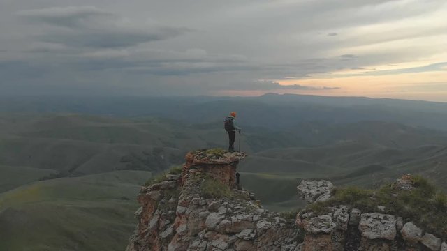 Aerial - Moving around a young adult woman with a backpack reaching the top of the rock at the foot of the epic plateau at sunset. View from the back. Russia. North Caucasus