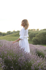 Fototapeta na wymiar attractive woman in white dress walking in violet lavender field and touching flowers