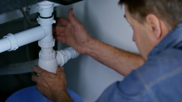 Tilt up of male plumber unscrewing drain pipe under sink and cleaning it while talking to unrecognizable female homeowner