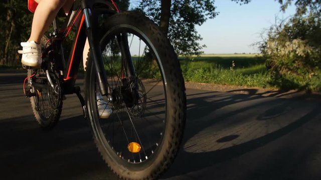 Closeup shot of teen girl legs riding bicycle on countryside road. Bike wheels spinning as teenage female cycling on rural road at sunset. Steadicam shot