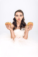 attractive young bride in wedding dress holding burgers in hands while grimacing and looking at camera isolated on white