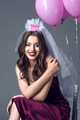 Obraz na płótnie Canvas happy future bride in veil for bachelorette party sitting on chair with tied pink balloons isolated on grey