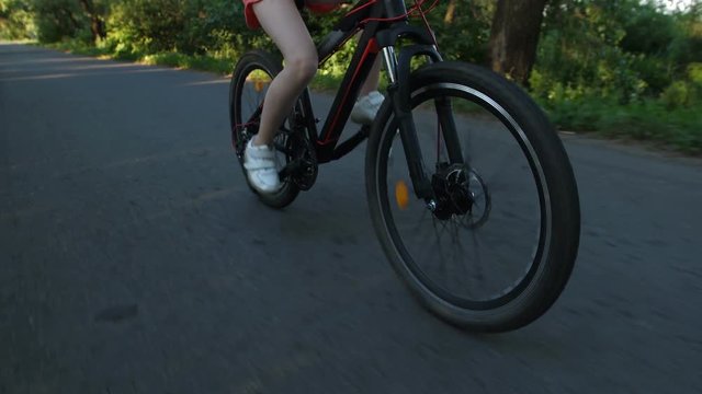 Closeup of teenage girl legs cycling on bicycle outdoors in countryside. Young female enjoying sunset and nature while riding bike on rural road. Steadicam shot