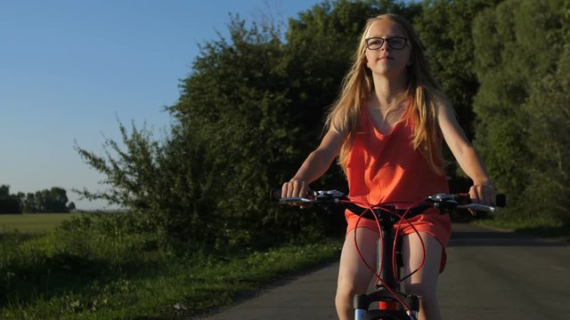 Portrait of cute blonde teenage girl riding bicycle and enjoying nature views in countryside at sunset. Positive teenager cycling on a trip on rural road. Steadicam shot