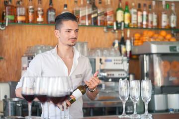 The barman is holding a bottle of red wine. The barman examines a bottle of red wine.The bartender pours red wine to the client in the hotel bar. The concept of service. Concentrate on the bartender.