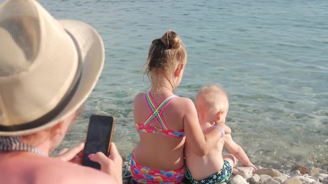 Happy mother taking picture of her children with smartphone on the beach outdoors. Mother taking picture of her family at the sea. Vacation concept.