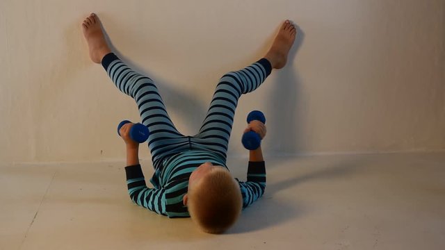 Happy child in striped pajamas is engaged in gymnastics with dumbbells at home.