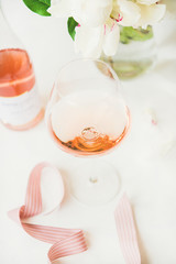 Rose wine in glass and bottle, pink decorative ribbon, peony flowers over white background, selective focus. Summer celebration, wedding greeting card, invitation concept