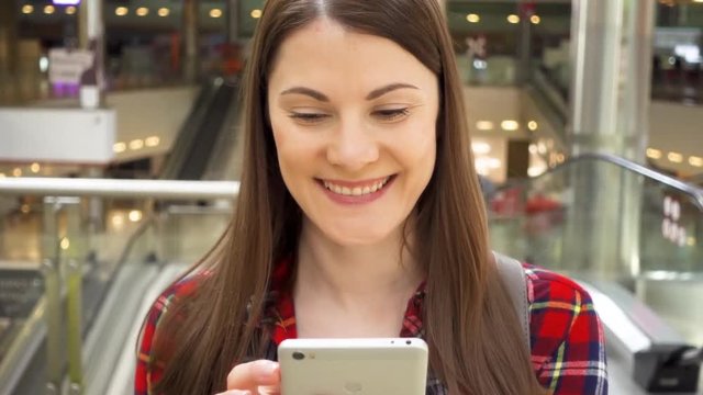 Young woman walking in shopping mall smiling in slow motion. Female using her smart phone, talking with friends via messenger app on mobile phone. Shopping consumerism concept