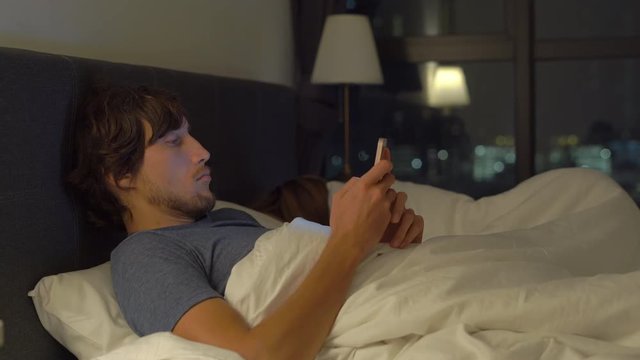 man and woman lying in bed, man with a smartphone while woman is sleeping. problems in marriage and intersexual relationship problems and mobile devices addiction concept